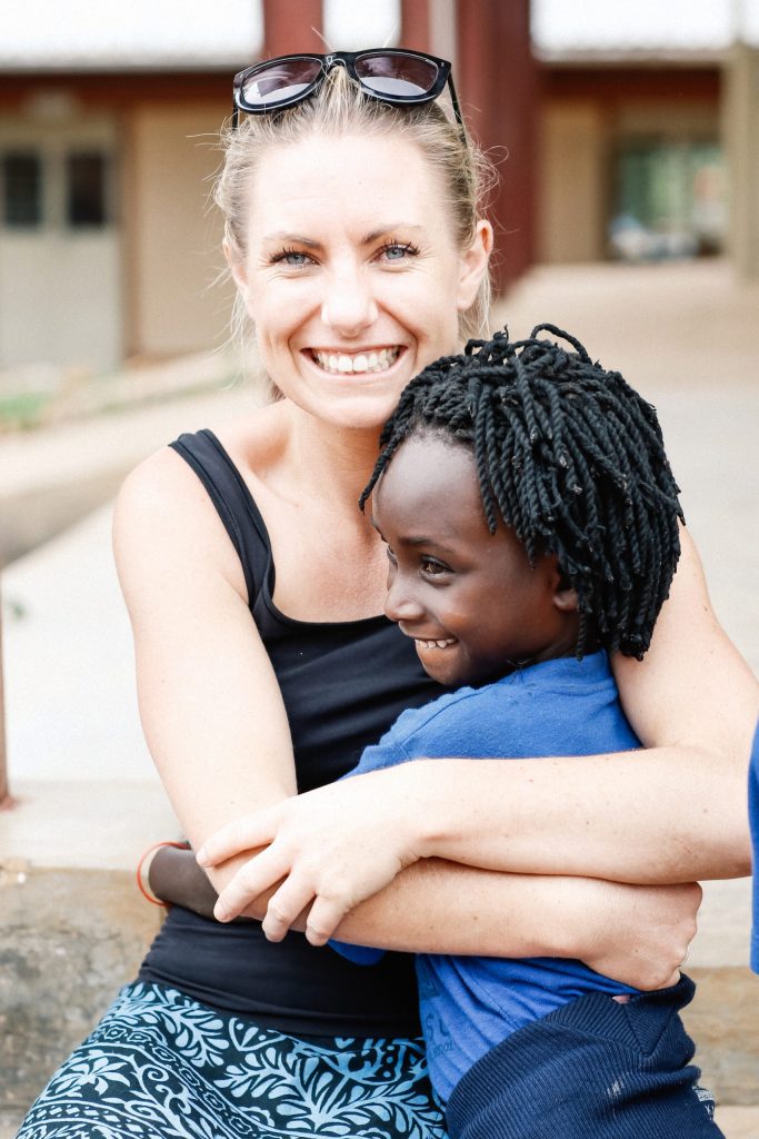 A smiling woman with sunglasses on her head embraces a joyful child. Both are outdoors, with the woman wearing a black tank top and patterned pants, embodying the essence of women in business and the balance they strike. The child, dressed in a blue shirt, stands against a backdrop of a building and some greenery.