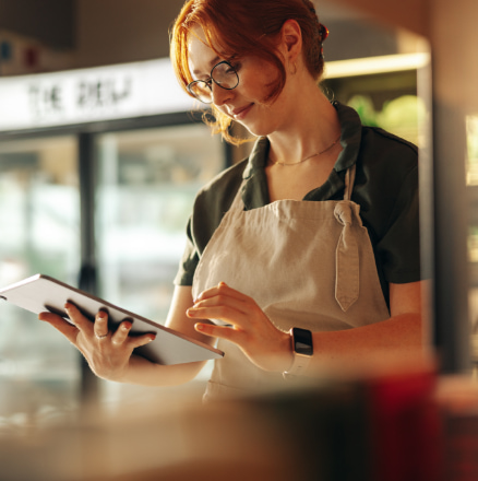 A woman with red hair and glasses stands in a cafe, wearing an apron and holding a tablet. She appears to be taking notes or entering information, embodying the spirit of women in business. Various shelves and items are visible in the background, showcasing her entrepreneurial environment.