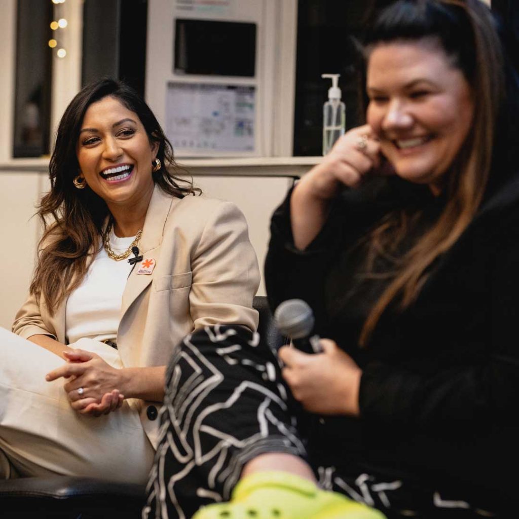 Two women are seated and engaged in a lively conversation during an indoor event. The woman on the left is wearing a beige blazer and smiling, while the woman on the right, holding a microphone, is laughing. They appear to be enjoying their discussion about business ideas for women entrepreneurs.