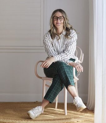 A person with long, wavy hair, glasses, and a smiling expression is sitting on a light pink chair in a brightly lit room. They are wearing a white, striped long-sleeve shirt, green plaid pants, and white sneakers. The background shows a white wall with a window and closed blinds—perfect for brainstorming business ideas for women.