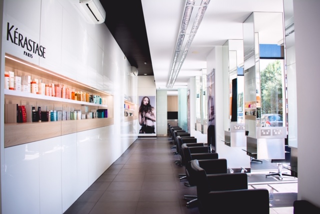 A modern, sleek hair salon with black chairs lining a white counter. Shelves stocked with hair products are on the left, under a "Kérastase Paris" sign. Large windows and mirrors reflect natural light, giving the space a bright, airy feel—an inspiring setting for women in business starting a venture.