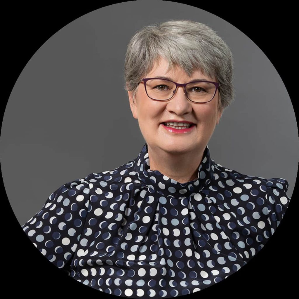 A person with short gray hair and glasses smiles at the camera. They are wearing a dark-colored shirt with a white polka dot pattern, against a gray background. This confident entrepreneur embodies the spirit of successful female businesses.