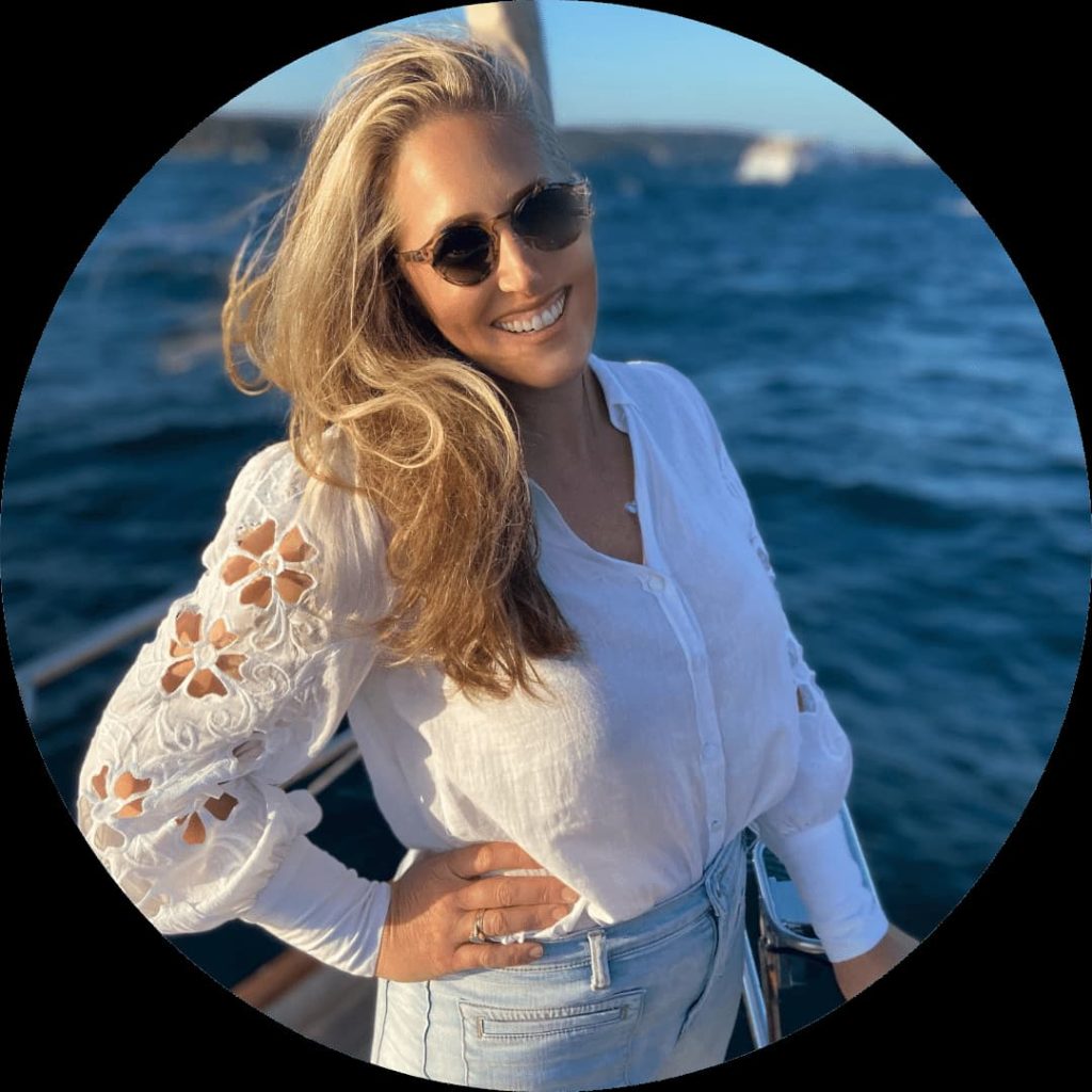 A person with long blonde hair is standing on a boat, smiling at the camera. They are wearing sunglasses, a white blouse with embroidered floral designs on the sleeves, and light blue jeans. The water and a distant shoreline are visible in the background—an inspiring image for starting a business.