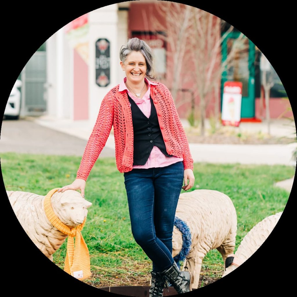 A person with short hair, wearing a pink shirt, black vest, jeans, and a coral cardigan, leans on the back of a sheep statue adorned with knitted accessories. This charming outdoor scene with buildings and greenery in the background exemplifies creative business ideas for women.