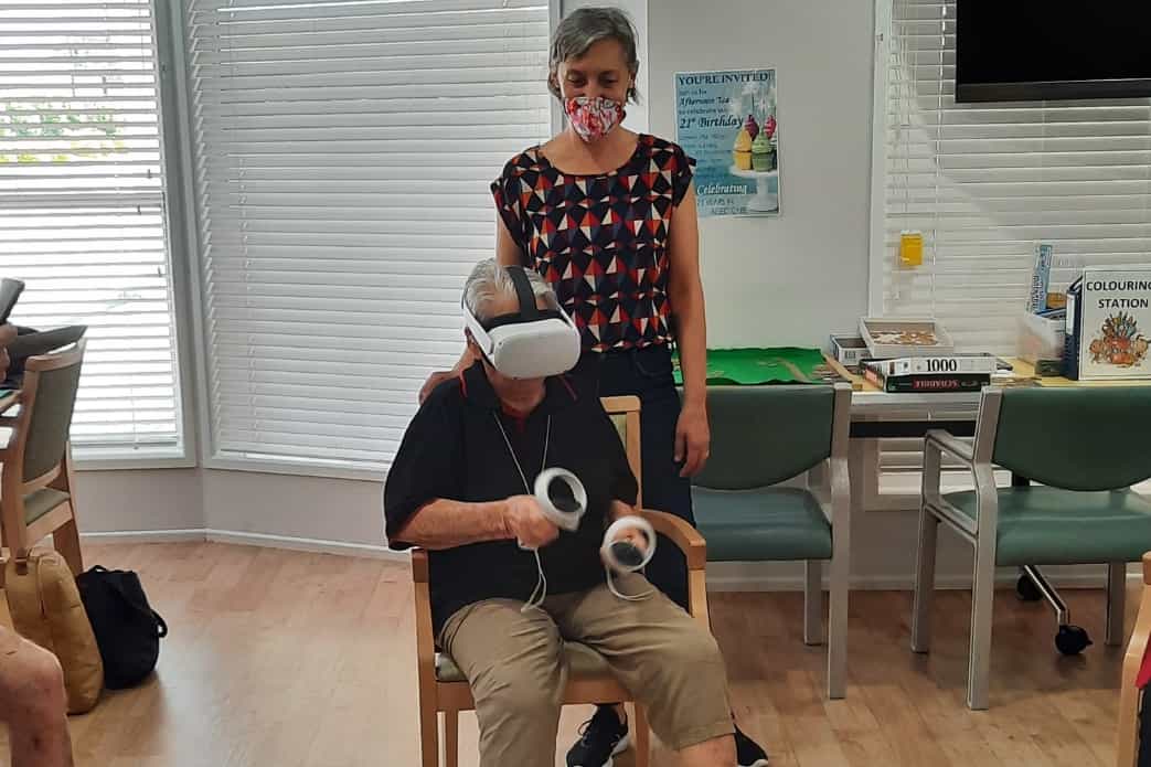 An elderly person is sitting in a chair using a VR headset and controllers, while a woman stands next to them, assisting. The setting appears to be a communal room with tables, chairs, and various activity supplies. Both individuals are wearing face masks. It’s inspiring to see how technology can foster connections in female businesses focused on seniors' well-being.