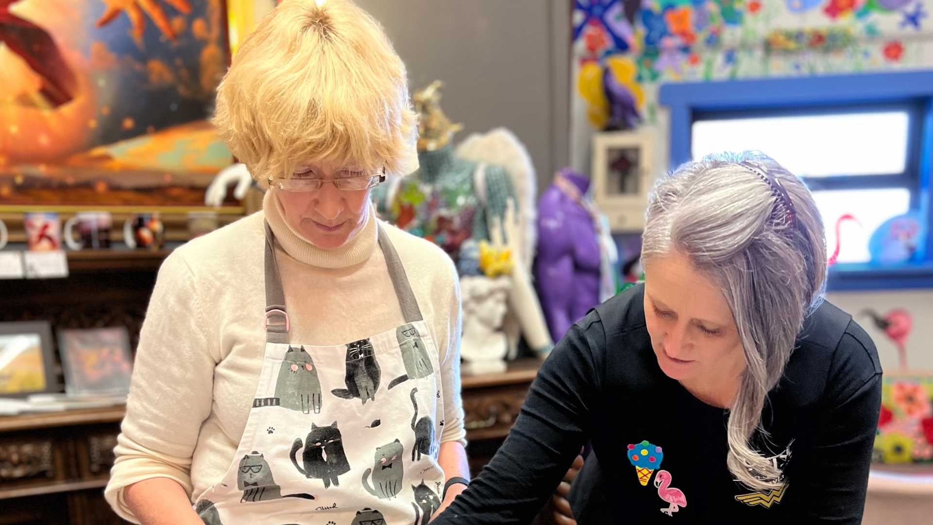 Two women are working together on an art project in a colorful room filled with various artwork and decorations. One woman, with short blonde hair, wears a cat-themed apron, while the other, with long gray hair, is focused on her task. Discussing business ideas for women, they chat about starting a business based on their creativity.