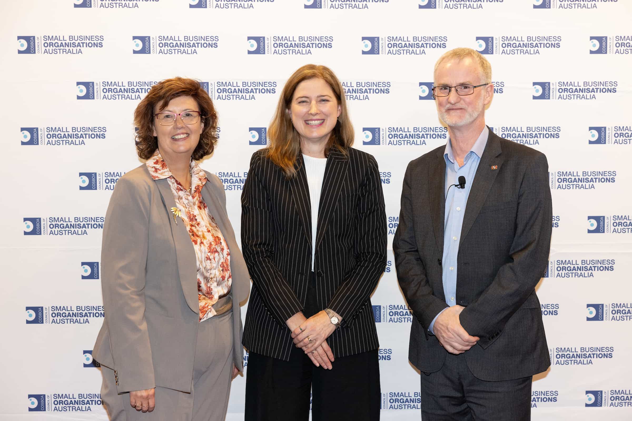 Three people are standing in front of a backdrop with the logo and text "Small Business Organisations Australia." They are smiling and dressed in business attire. The woman on the left wears a beige suit, the woman in the middle wears a black suit, and both exemplify successful women in business as they stand with their male colleague.
