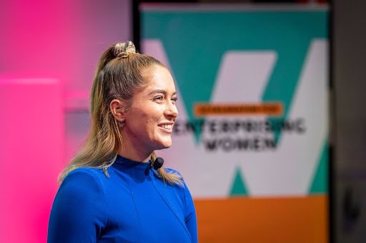 A person with long blonde hair in a ponytail, wearing a blue top, is speaking in front of a colorful backdrop. The backdrop features the text "EMPOWERING WOMEN" and has a large white letter "W" on a teal and orange background, inspiring business ideas for women to excel in starting a business.