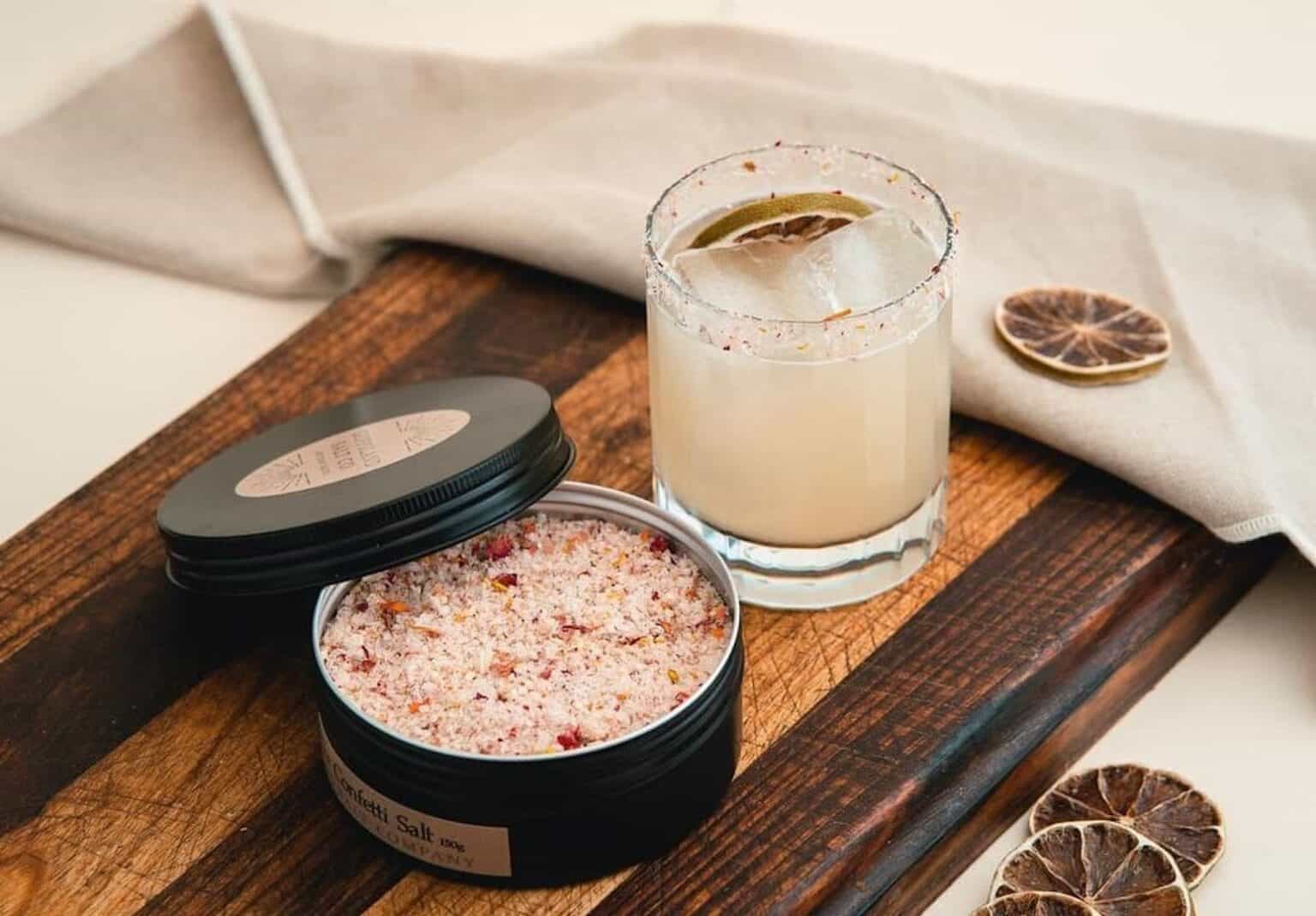 A glass of margarita with a salted rim, garnished with a slice of dried lime, sits on a wooden serving board next to an open tin of flavored salt—a simple yet elegant setup perfect for an entrepreneur starting a business. A beige napkin is folded in the background, and two additional slices of dried lime rest on the board.