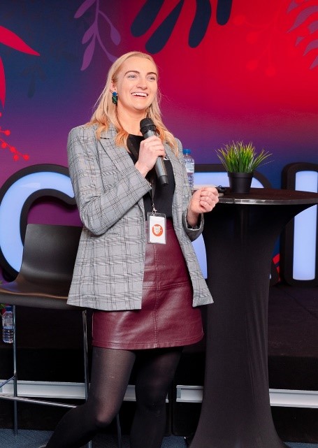 A person with long blond hair stands on a stage holding a microphone. They are wearing a gray checkered blazer, a maroon leather skirt, black tights, and a lanyard with an ID. A small green plant and water bottle are on the round black table next to them. The background features colorful abstract designs showcasing women in business.