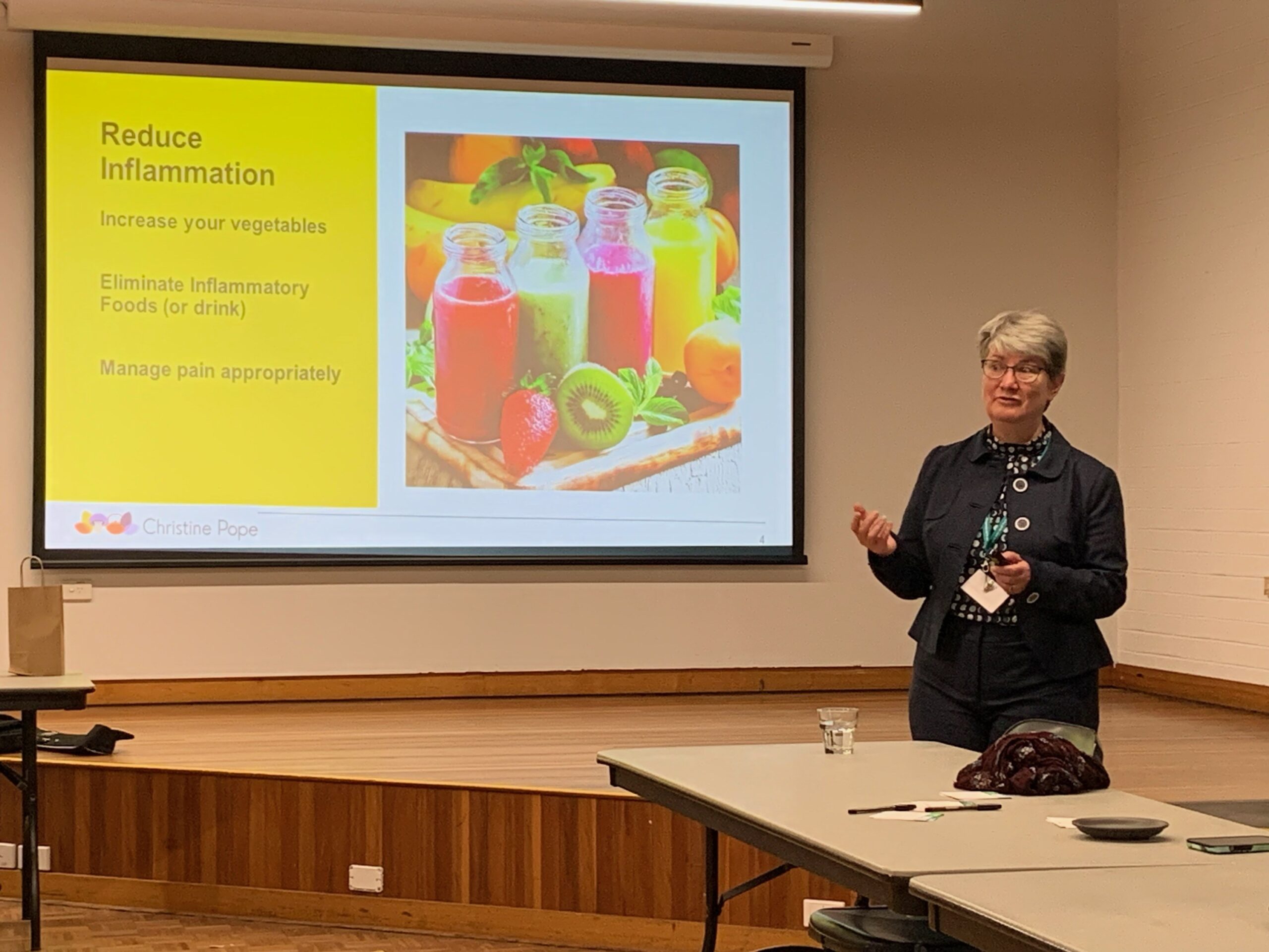 A woman in business stands next to a projector screen during a presentation. The screen displays a slide titled "Reduce Inflammation," featuring colorful smoothie bottles, fruits, and text about increasing vegetables, eliminating inflammatory foods, and managing pain appropriately.