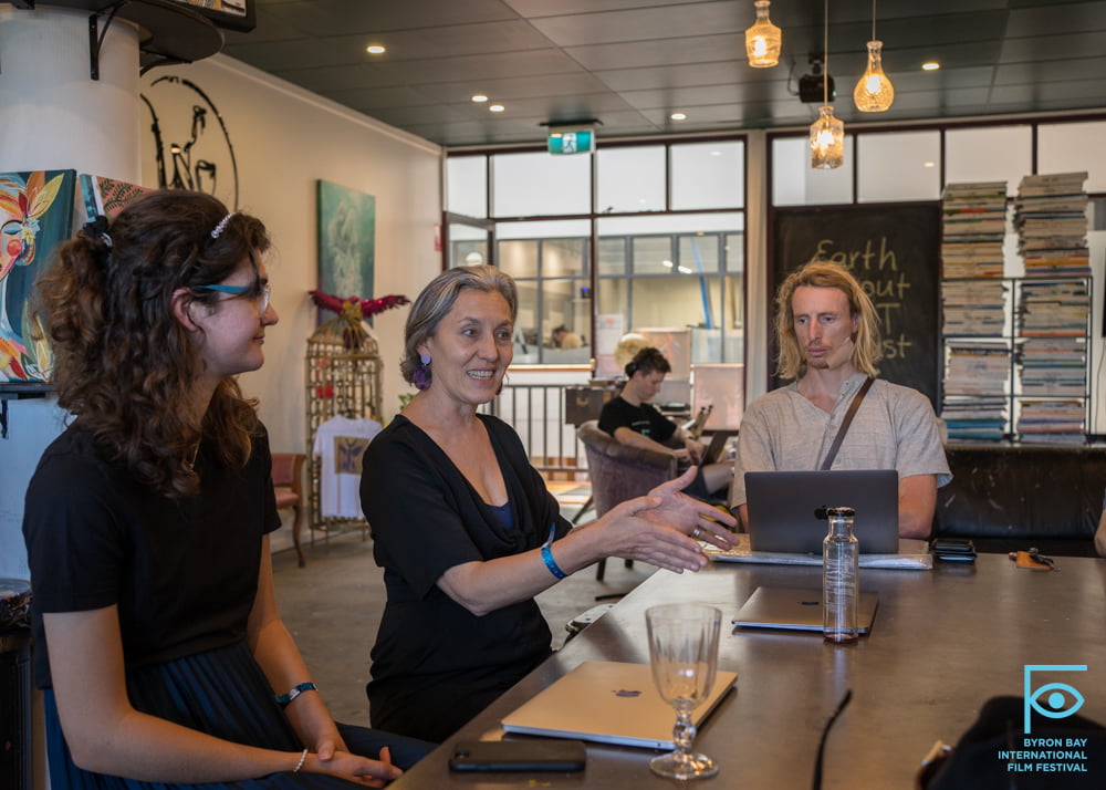 Three people are sitting at a table in a café. The middle person, an entrepreneur, is speaking and gesturing with her hands. The person on the left is smiling while the one on the right is using a laptop. A “Byron Bay International Film Festival” sign is visible in the lower right corner.