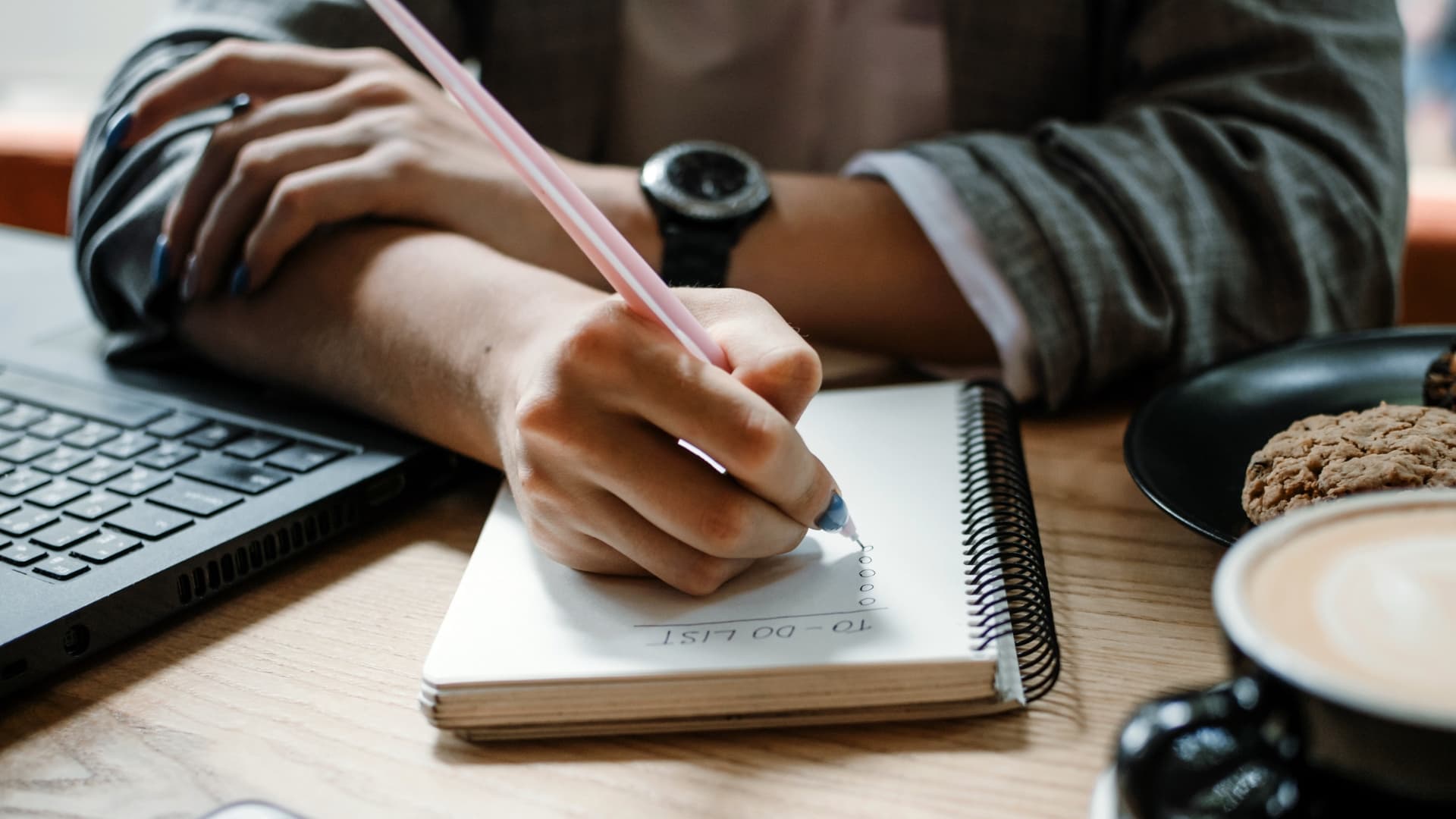 A person, possibly an entrepreneur starting a business, is writing a to-do list in a spiral notebook with a pink pen while seated at a wooden table. A watch is visible on their wrist. Nearby are a laptop, a plate with cookies, and a cup of coffee.