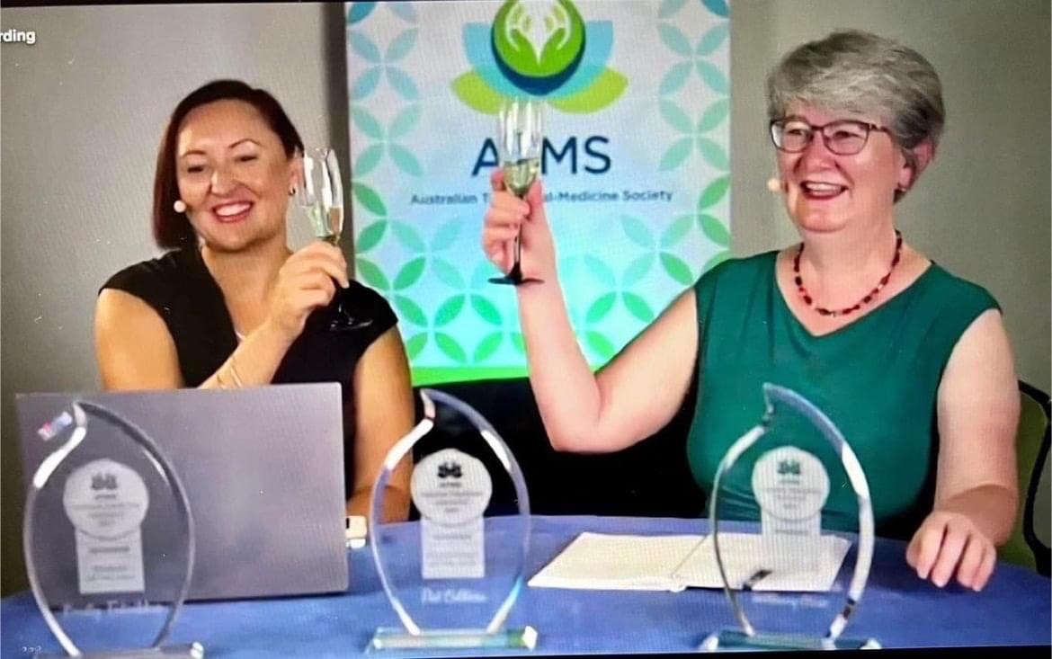 Two women in business smile and raise glasses in a toast in front of an AMS banner. They sit at a table with three trophies and a laptop, celebrating entrepreneurial success. The woman on the left wears a black dress, while the woman on the right sports a grey dress with a red necklace.