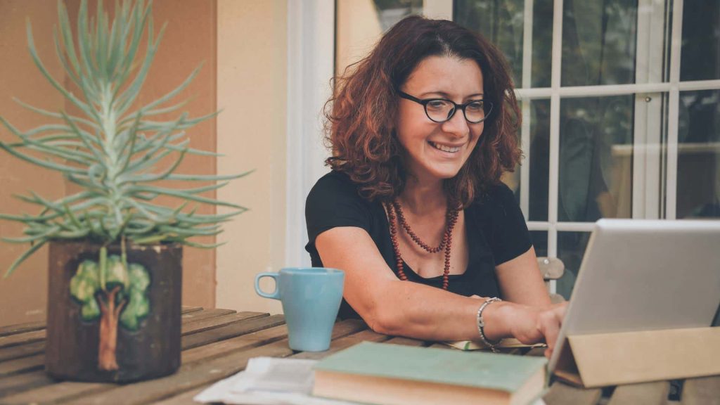 A woman with curly hair and glasses smiles while using a tablet at a wooden table. She sits outdoors with a large plant, a blue mug, and books on the table in front of her, researching business ideas for women. A glass door is in the background.