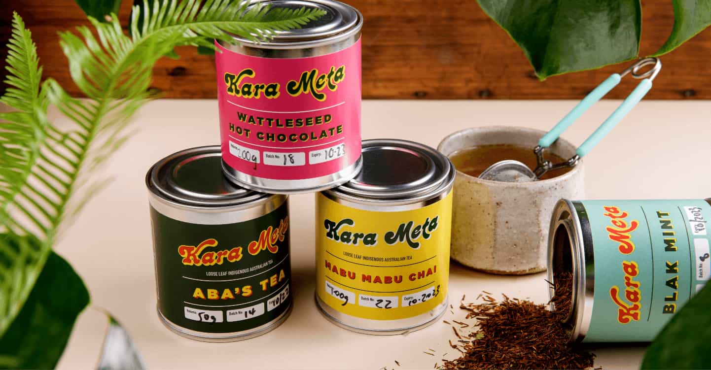Four colorful cans of tea and hot chocolate, labeled "Kara Meta," are displayed on a table. One can is open, spilling leaves, and a spoon is in a mug nearby. A plant is in the foreground, while more greenery decorates the background, showcasing perfect business ideas for women entrepreneurs.