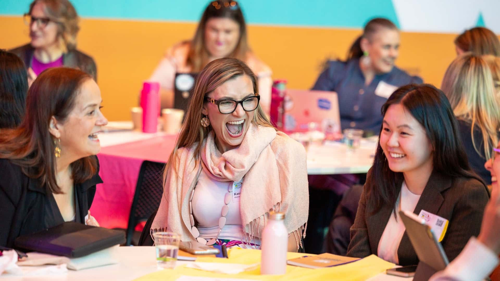A group of women in business sit together at a table, chatting and laughing during a conference or workshop. They are surrounded by papers, laptops, and drinks, indicating an engaging and collaborative environment. The background shows other participants involved in similar activities.