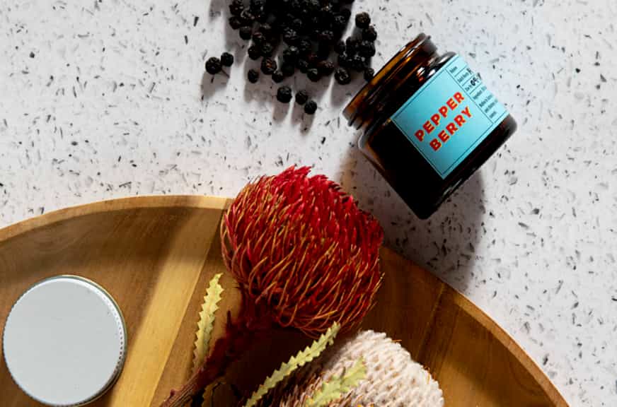 A small jar labeled "Pepper Berry" lies on a speckled white surface with its lid off, next to a scattering of pepper berries. Below it, a wooden plate holds two red and white botanical flowers and a small silver lid—a perfect setup for any women in business looking to add charm and flavor while starting a business.