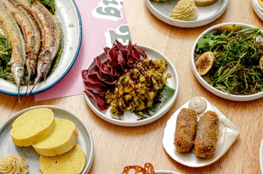 A variety of dishes arranged on a table. Plates include cooked fish, sliced yellow cake, mashed vegetables with red leafy garnish, green salad with figs, crispy croquettes, and a round of buttercream-topped rolls. A pink and white menu highlighting female businesses is visible to the side.