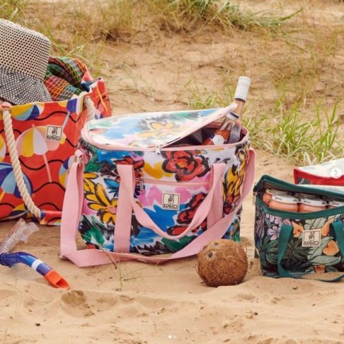 A colorful beach setup featuring three vibrant, floral-patterned cooler bags on the sand, showcasing perfect business ideas for women. One bag has ropes for handles, another has a pink zipper with a wine bottle peeking out, and the third holds cans. An open coconut and beach grass complete the scene.