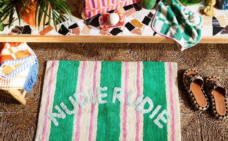 A colorful scene with a green, pink, and white striped rug that reads "NUDIE RUDIE" placed on a woven mat. Nearby are patterned towels, a fruit bowl, a plant, and leopard-print sandals. The backdrop has a mosaic countertop with various colorful items—a perfect setting to inspire business ideas for women.