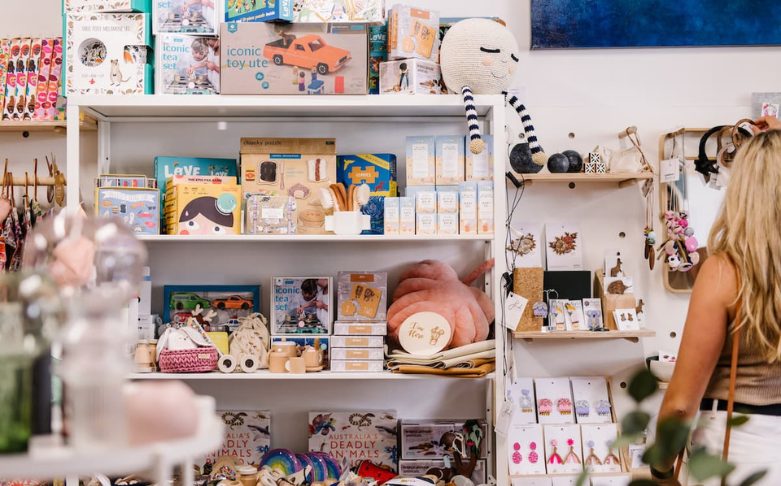 A woman with blonde hair browses products in a colorful store. The shelves are filled with various toys, books, and gift items, including stuffed animals, educational kits, and decorative pieces. The ambiance is bright and inviting—a perfect business idea for aspiring women entrepreneurs.