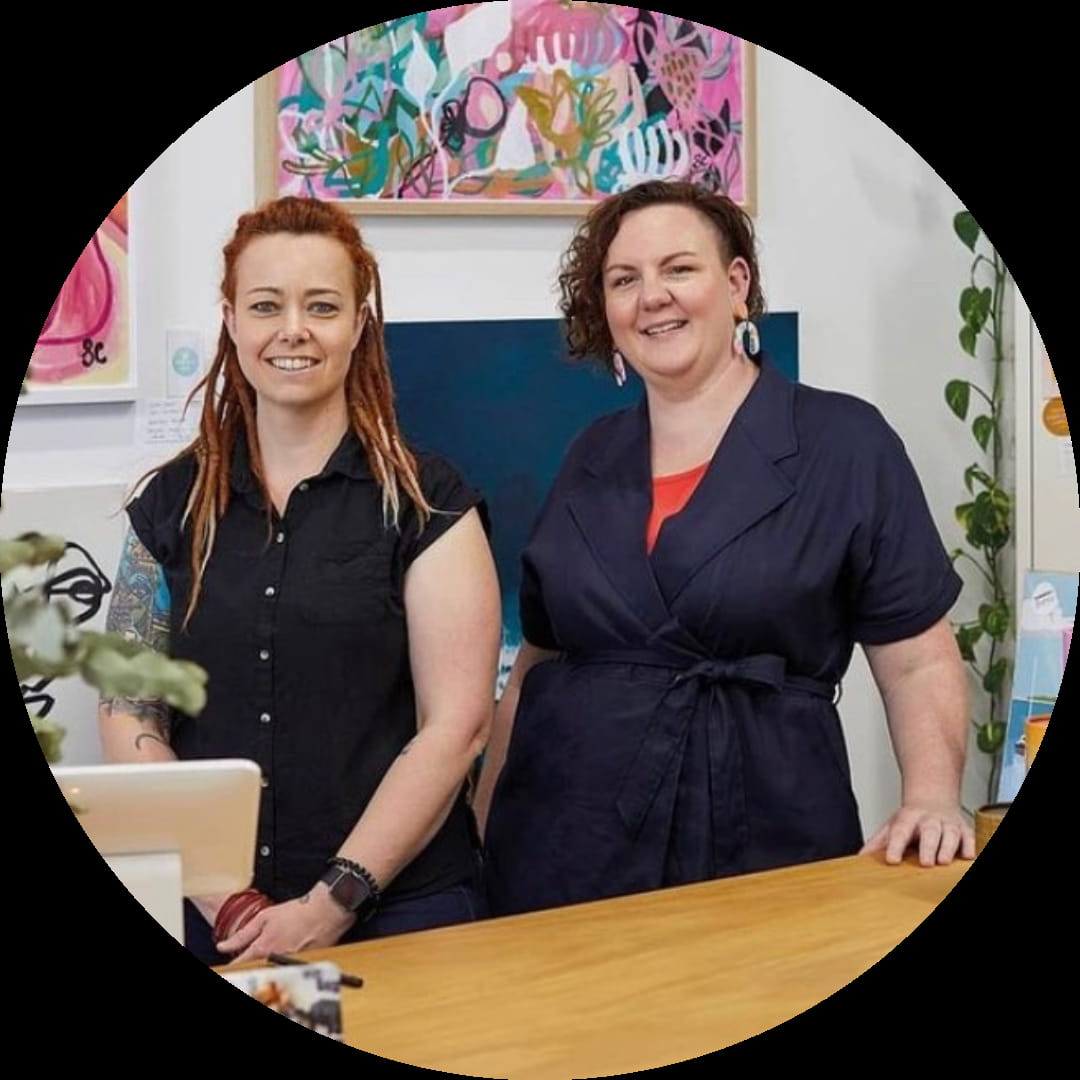 Two entrepreneurs smile while standing behind a counter in an art gallery. Both wear casual clothing: a black sleeveless shirt and a black short-sleeved jacket. Bright paintings with floral and abstract designs adorn the wall behind them, showcasing their creativity and passion as women in business.