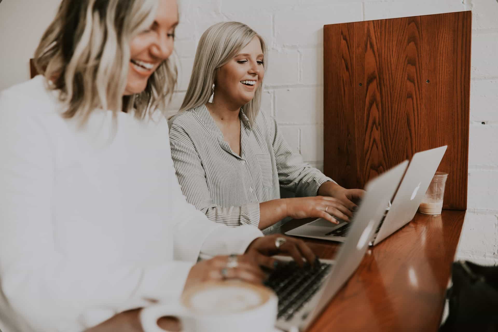 Two women in business are sitting at a wooden table with laptops in front of them, smiling and typing. One has a coffee cup in the foreground. The background features a white brick wall and wooden panel. They seem to be enjoying their work or conversation, perhaps discussing ideas on starting a business.
