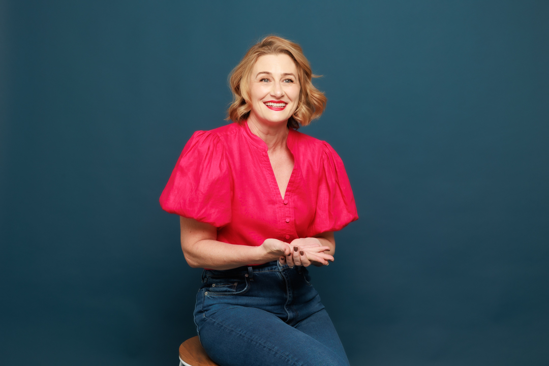 A smiling entrepreneur with shoulder-length blonde hair is sitting on a stool against a dark blue background. She is wearing a bright pink blouse with puffed sleeves and high-waisted blue jeans. Her hands are gesturing gently in front of her, embodying the spirit of women in business.