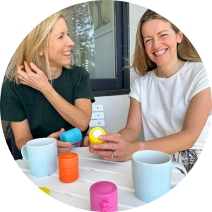 Two women sitting at a table adorned with colorful small containers and white mugs, smiling and engaged in conversation. One woman, with shoulder-length blonde hair, is wearing a dark green shirt and the other, with light brown hair, is in a white shirt. Both are successful entrepreneurs discussing their enterprises.