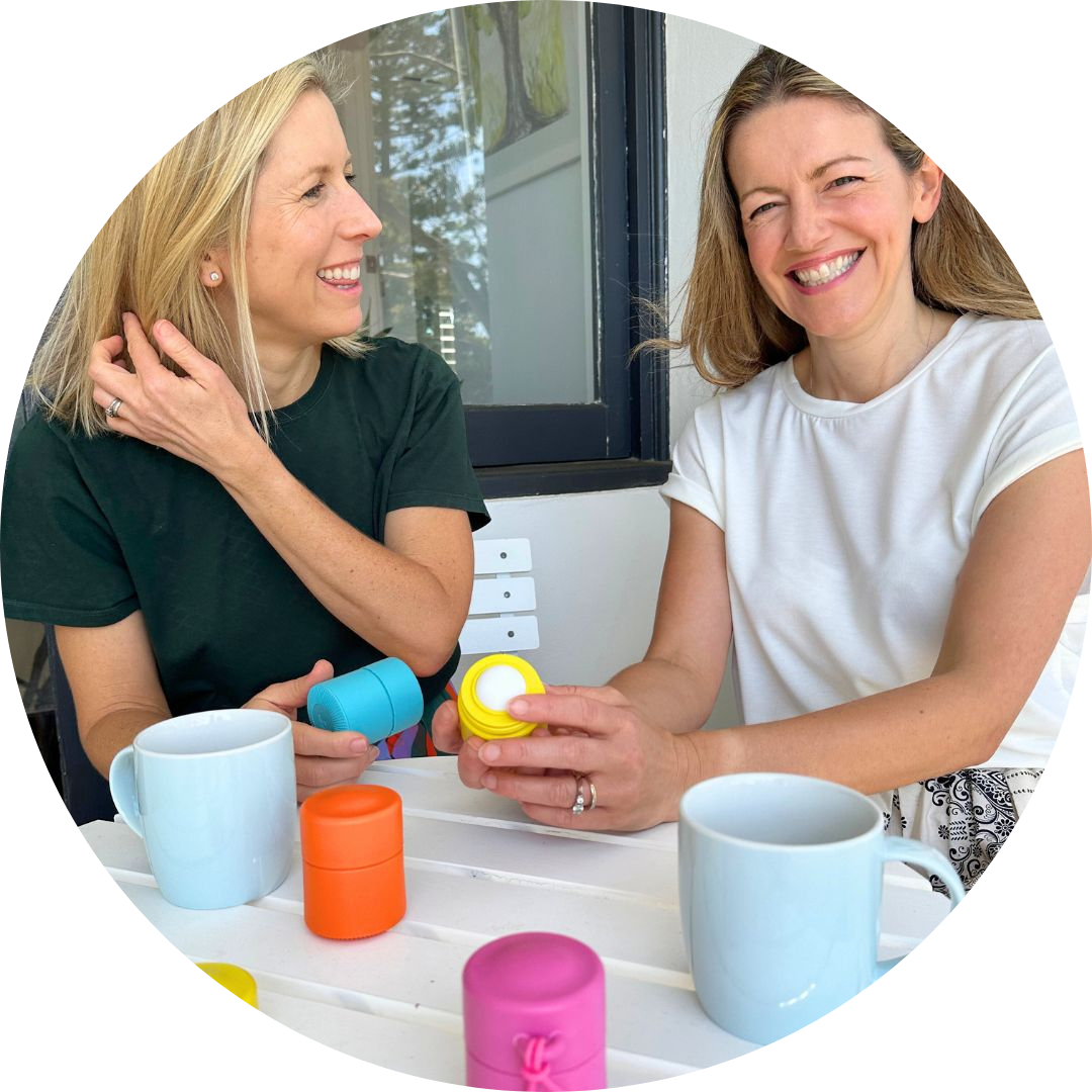 Two women sitting at a table adorned with colorful small containers and white mugs, smiling and engaged in conversation. One woman, with shoulder-length blonde hair, is wearing a dark green shirt and the other, with light brown hair, is in a white shirt. Both are successful entrepreneurs discussing their enterprises.