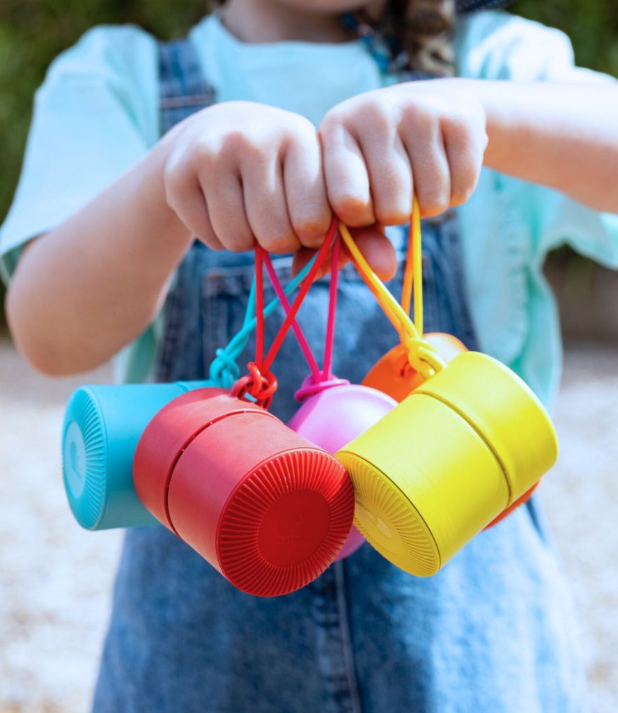A child is holding colorful, cylindrical containers with handles, perhaps dreaming of starting a business. The child is wearing a light blue shirt and denim overalls. The handles of the containers are looped around the child's fingers, and the containers are red, yellow, blue, and pink.