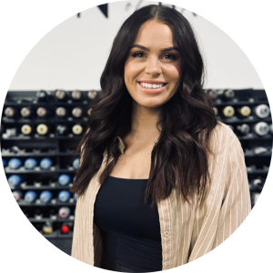 A woman with long, dark hair wearing a black top and a light-colored, striped shirt smiles at the camera. She is standing in front of shelving filled with rows of organized spools or rolls, creating an industrial or craft-like background that inspires business ideas for women entrepreneurs.