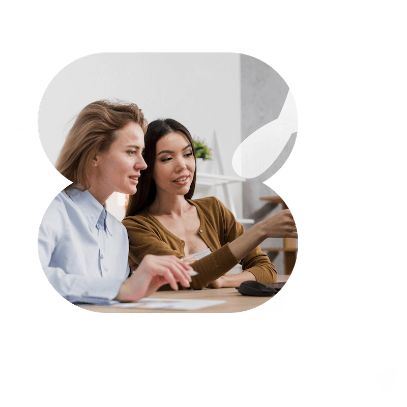 Two women sitting at a table, focused on a computer screen. One points at the screen while the other holds a pencil. The image is framed with abstract black and white shapes in the corners.