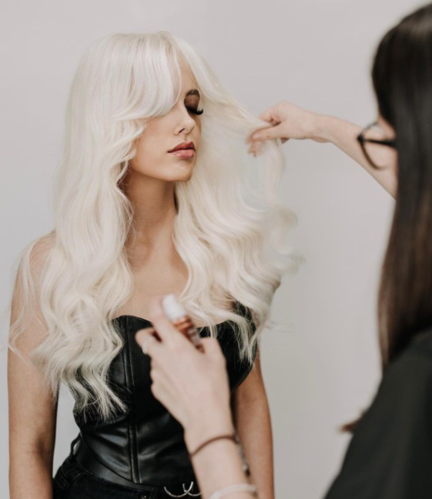 A woman with long, wavy platinum blonde hair is having her hair styled by a hairdresser. She is wearing a black, strapless top and has her eyes closed. The hairdresser, considering business ideas for women, holds a section of the woman's hair and a spray can in the other hand.