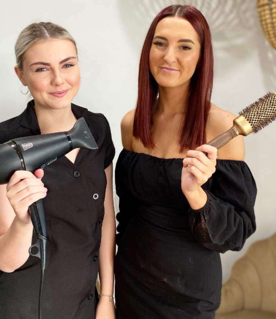 Two women wearing black clothes stand smiling. The woman on the left holds a hair dryer, and the woman on the right holds a round hairbrush. They are likely entrepreneurs, posing confidently in a well-lit salon setting—a great example of successful female businesses in action.