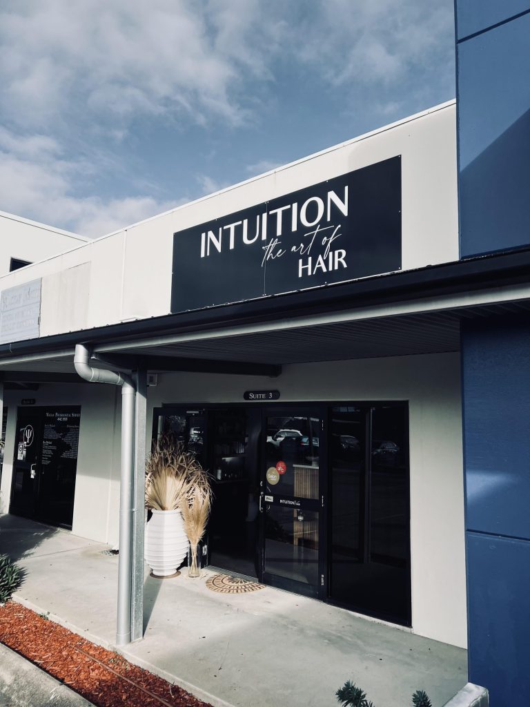 A storefront with a sign reading "INTUITION the art of HAIR." The exterior features gray and blue panels, a large planter with dried decorative grass, and glass double doors leading into the salon. Perfect for women in business seeking inspiration, the sky above is partly cloudy.