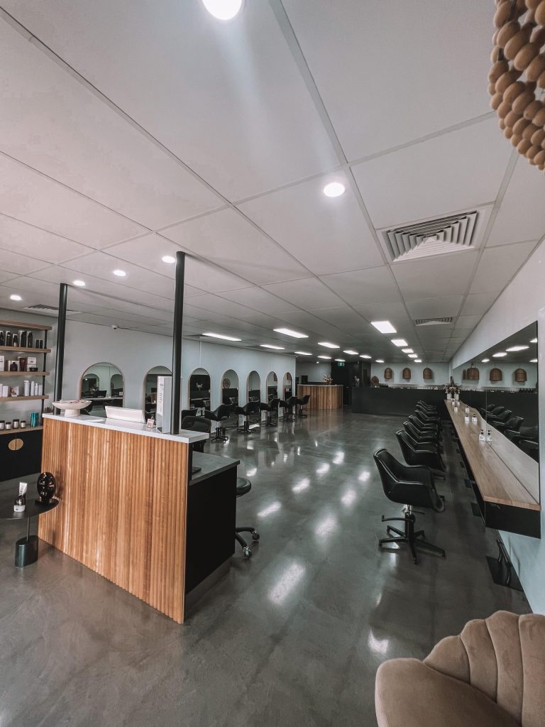 A modern and spacious hair salon with a sleek design, perfect for aspiring female entrepreneurs. On the left, there's a wooden reception desk and product shelves. The right side features a long line of styling chairs and mirrors. Bright ceiling lights illuminate the polished concrete floor.
