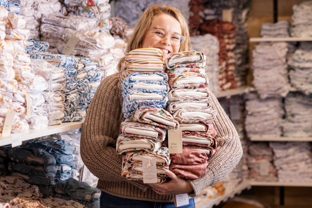 A person with glasses and blonde hair smiles while holding a large stack of folded fabrics of various colors and patterns. They are surrounded by shelves filled with neatly arranged, similar-looking folded fabrics in what appears to be a fabric store—an inspiring example of successful female businesses.