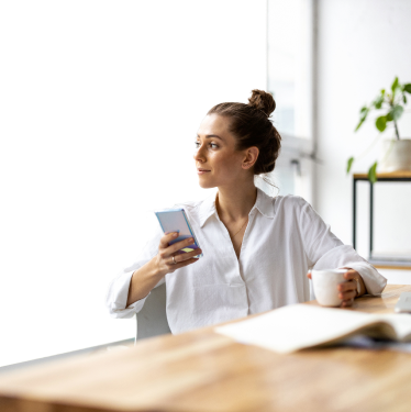 A woman with her hair in a bun, wearing a white shirt, holds a smartphone in one hand and a cup in the other while sitting at a table with an open book. She is looking to the side, with a potted plant and window in the background—an entrepreneur envisioning her next move.