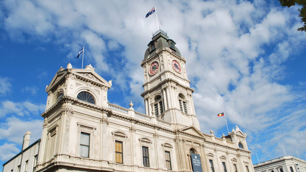 The image depicts a historic town hall building with a prominent clock tower and ornate architectural details. The building features two flags flying on its rooftop: a national flag and another flag. Imagine the inspiring backdrop this could provide for sharing innovative business ideas for women. The sky above is blue with scattered white clouds.