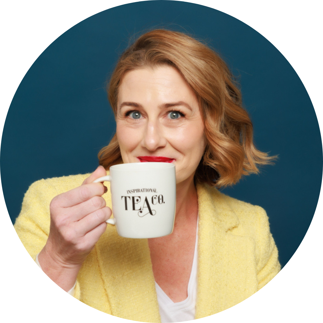 A person with shoulder-length blonde hair and red lipstick smiles while holding a white mug with the words "Inspirational Tea Co." They are wearing a yellow blazer and a white top, against a dark blue background. This scene exudes the energy of an entrepreneur starting a business focused on creative business ideas for women.