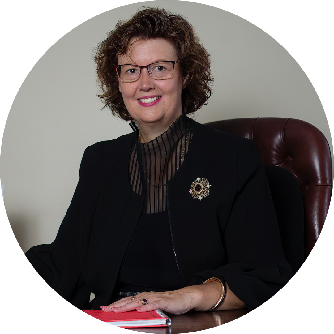 A woman with short curly brown hair and glasses sits at a desk. Dressed in a black outfit with a decorative brooch and sheer neckline, she holds a red book. Smiling confidently, she embodies the spirit of women in business. An office chair is visible in the background.