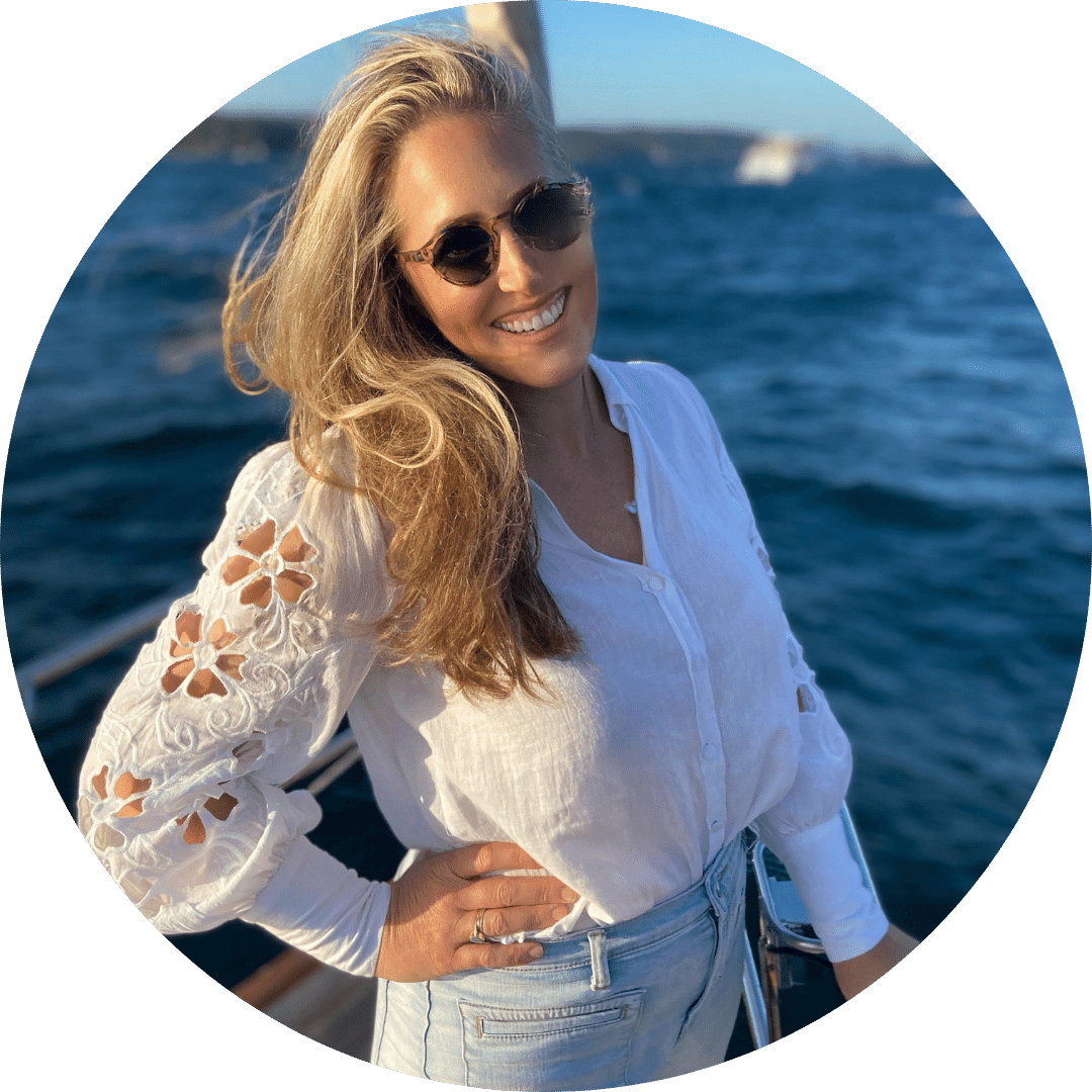 A woman with long blonde hair is smiling and wearing sunglasses. She is standing on a boat with her hand on her hip, dressed in a white blouse with lace details and light blue jeans. The background features a body of water and a distant shoreline, capturing the essence of inspiration for female businesses.