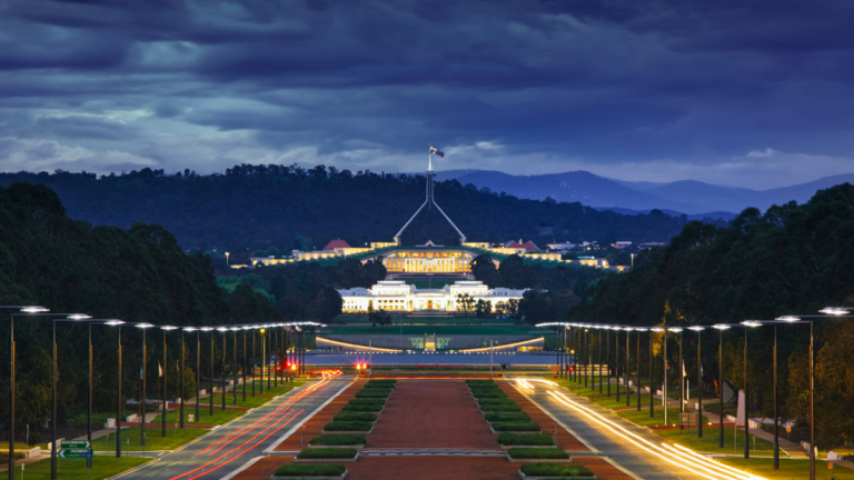 A twilight view of Canberra's Parliament House, featuring the iconic flagpole centered on the roof. Illuminated buildings and well-lit streets are prominent in the foreground, with motion blur from vehicle lights. Dark trees and rolling hills create a scenic backdrop, inspiring entrepreneurial visions for female businesses.