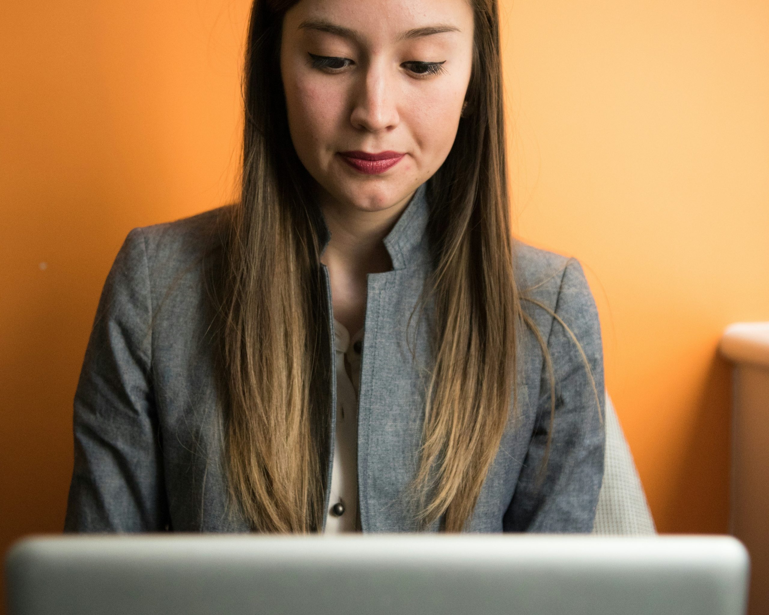 A person with long, straight hair is sitting in front of a laptop. Wearing a gray blazer and a light-colored shirt, they appear focused on the screen, possibly contemplating new business ideas for women. The background is a solid orange color.