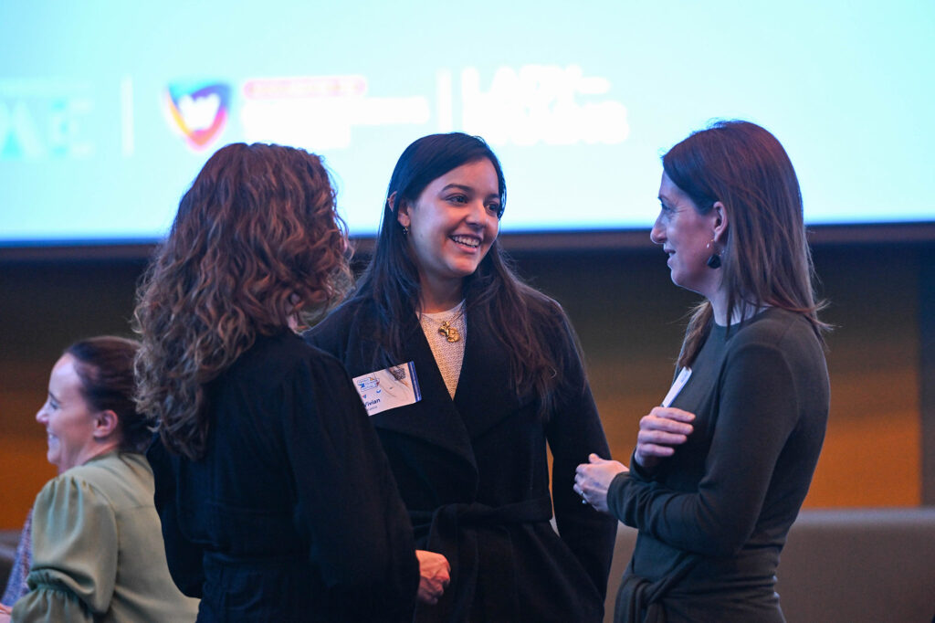 Four women are engaged in a lively conversation at a networking event. Three of them are standing and smiling at each other while the fourth woman, an aspiring entrepreneur, sits slightly apart, smiling. A large screen in the background displays some logos and text about business ideas for women.