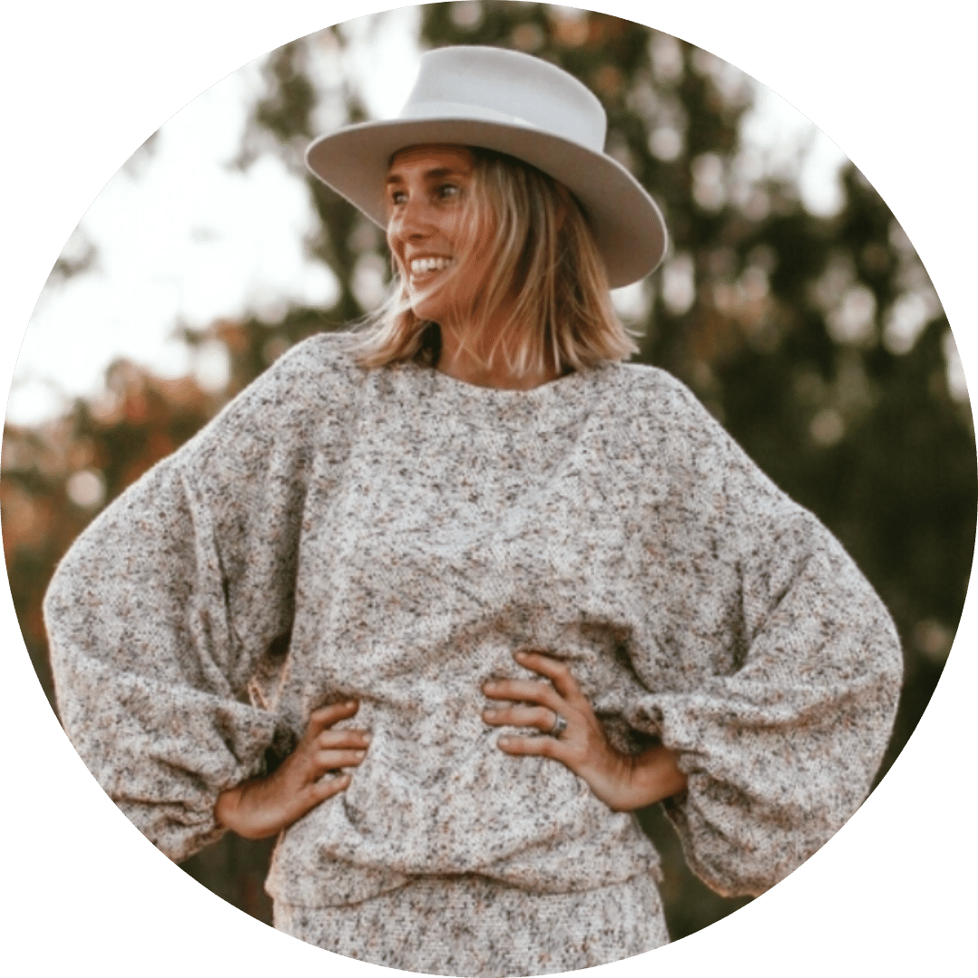 A person with chin-length blonde hair is smiling and looking to the side, perhaps contemplating new business ideas for women. They are wearing a wide-brimmed hat and a cozy, loose-fitting knit sweater. The background is blurred with trees and foliage.