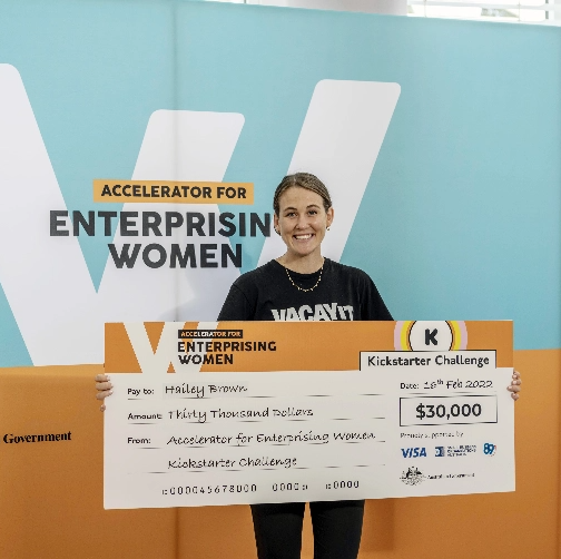 A woman holds a large check for $30,000 awarded for the Kickstarter Challenge, dated February 18, 2022. She stands in front of a banner that reads "Accelerator for Enterprising Women," celebrating her success in starting a business and smiles at the camera.