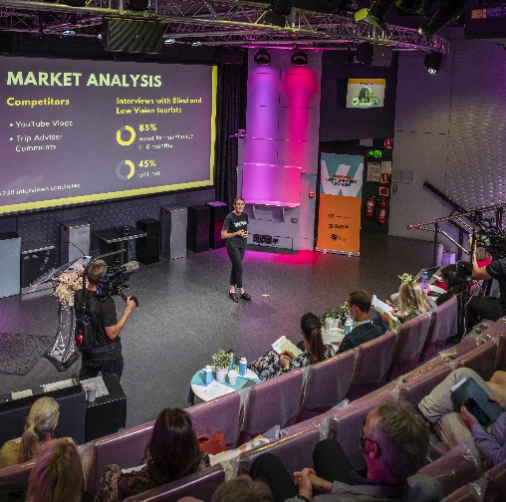 A presenter stands on stage in front of a large screen displaying a market analysis slide. The audience, filled with aspiring entrepreneurs, watches attentively in a modern venue with pink and purple lighting. The screen shows data on competitors and user surveys. A camera operator films the presentation.